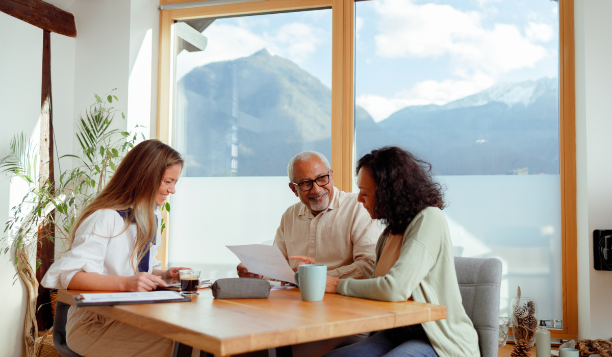 A woman, likely a financial advisor, is seated with a senior couple in a bright room with a stunning mountain view, indicative of a home in Slovenia.